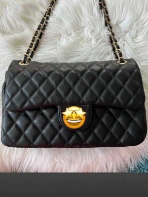 Svart quiltad handväska med kedja - Stilren svart handväska med quiltat mönster och kedjedetaljer på axelbanden. Väskan har en klassisk rektangulär form och elegant lock med metallspänne. Perfekt för dig som gillar tidlös design och lyxig känsla.
