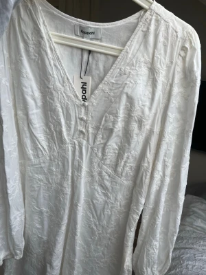 Vit studentklänning  - Vit kortklänning från Kappahl med v-ringning och långa ärmar som liknar Adoors populära klänning. Klänningen har ett diskret blommigt broderat mönster. 