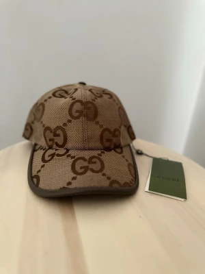 Beige keps med GG-mönster från Gucci - Snygg beige keps från Gucci med klassiskt GG-logomönster i brunt. Kepsen har böjd skärm och är tillverkad i ett slitstarkt tygmaterial. Perfekt för dig som vill ha en trendig accessoar med lyxig känsla.