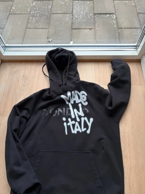 Svart Dondup hoodie med tryck - Säljer en svart hoodie från Dondup med stor huva och magficka. På bröstet står det 'MADE IN ITALY' i vit graffiti-stil och Dondup i svart. Hoodien har en avslappnad passform och är tillverkad i mjukt bomullsmaterial. Perfekt för en streetwear-look.