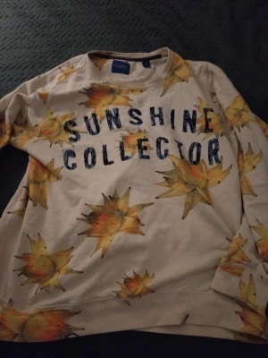 Scotch & Soda sweatshirt med print - Cool sweatshirt från Scotch & Soda med texten 'Sunshine Collector' och stora solrosliknande blommor i gult och orange på en ljus bakgrund. Rund halsringning och ribbade muddar. Perfekt för dig som gillar unika prints och vill sticka ut.