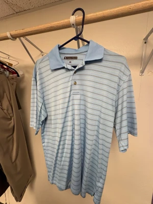 Ljusblå randig pikétröja Pebble Beach - Säljer en ljusblå pikétröja från Pebble Beach med tunna vita och grå ränder. Tröjan har klassisk krage, två knappar och korta ärmar. Materialet är performance-tyg som är lätt och bekvämt. Perfekt för en avslappnad och sportig look. Ord pris: ca 1000kr. Passar för L också.