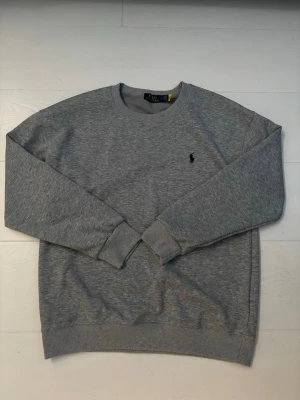 Grå sweatshirt från Polo Ralph Lauren - Klassisk grå sweatshirt från Polo Ralph Lauren med rund halsringning och broderad logga på bröstet. Tröjan har ribbade muddar vid ärmslut och nederkant. Perfekt för en avslappnad och stilren look. Tröjan passar bättre på M den är lite mindre i storlek