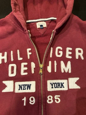 Röd Hilfiger Denim hoodie med dragkedja - Snygg röd hoodie från Hilfiger Denim med dragkedja, huva och stora vita tryck på bröstet där det står 'Hilfiger Denim New York 1985'. Mjuk insida och känguruficka framtill. Perfekt för en avslappnad streetstil.