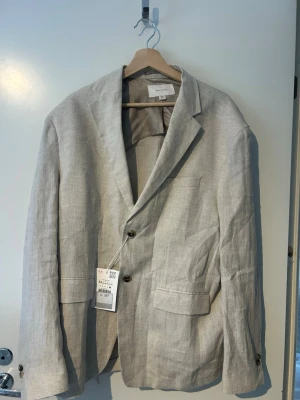 Beige linnékavaj från Zara Man - Stilren beige kavaj från Zara Man i luftigt linne. Klassisk passform med två knappar, tre framfickor och slits bak. Perfekt för en avslappnad men ändå dressad look. Snygga detaljer som hornknappar och mjuk axelstruktur.