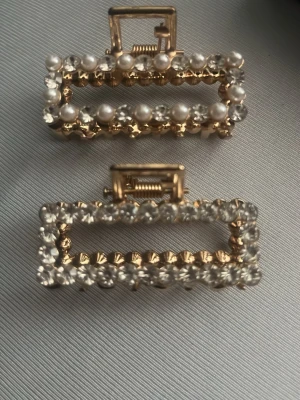 Guldiga hårklämmor med pärlor & stenar - Två rektangulära hårklämmor i guldfärgad metall, en dekorerad med vita pärlor och klara stenar, den andra med endast klara stenar. Perfekta för att ge frisyren en glittrig och elegant touch. Säljs som par.