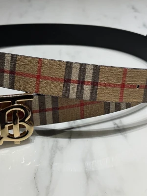 Burberry bälte med klassiskt rutmönster - Snyggt bälte från Burberry med det ikoniska rutmönstret i beige, svart, vitt och rött. Bältet har ett stilrent guldspänne med Burberrys logotyp. Perfekt accessoar för att lyfta din outfit.