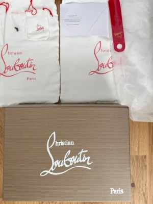 Svarta Louboutin sneakers med nitar - Säljer ett par svarta sneakers från Christian Louboutin med klassisk röd sula och svarta nitar på tån. Skorna har snörning, är tillverkade i mocka och har en lyxig vibe. Perfekta för dig som vill sticka ut med exklusiva detaljer och edgy look. 