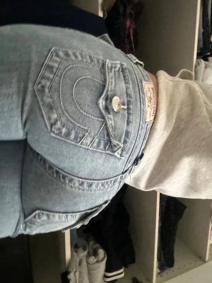 True Religion Joey ljusblå flare jeans - Säljer ett par ljusblå True Religion Joey jeans med låg midja och flare/bootcut passform. Klassiska kontrastsömmar, snygga fickdetaljer och silverfärgade knappar. Perfekta för dig som gillar en avslappnad men trendig look.