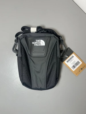 Svart axelväska från The North Face - Snygg svart axelväska från The North Face med justerbar rem och mesh-detaljer på framsidan. Väskan har logga både fram och på sidan, samt praktiska fack för småprylar. Tillverkad i slitstarkt syntetmaterial och perfekt för dig som vill ha en sportig och stilren look.  Modell: "Y2K SHOULDER BAG TNF BLACK-ASPHA" Mått: 20 × 15 × 7 cm