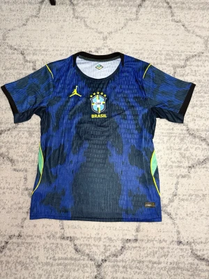 Blå Brasilien fotbollströja Nike - Snygg blå Brasilien fotbollströja från Nike med gul logga och CBF-emblem på bröstet. Tröjan har ett mönster i olika blå nyanser, svarta detaljer vid ärmslut och gula samt gröna inslag på sidorna. Perfekt för fotbollsträning eller att bära som supporter.