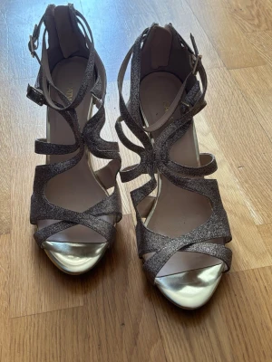 Glittriga sandaler med klack - Säljer ett par glittriga sandaler från Catwalk (Deichmann) med höga klackar och öppna tår. Skorna har flera smala remmar i glittrigt material och metallic guld på tån och klacken. Justerbart spänne vid ankeln för bästa passform.