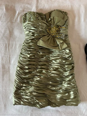 Grön axelbandslös kort klänning - Snygg axelbandslös kort klänning i glansigt grönt tyg med draperad effekt. Överdelen är dekorerad med pärlor och paljetter samt en stor rosett framtill med glittrande detaljer. Perfekt för dig som vill sticka ut.