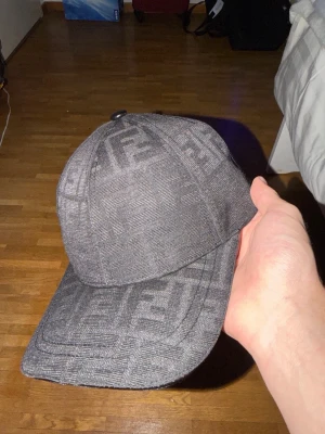 Grå Fendi keps med FF-mönster - Snygg grå keps från Fendi med det klassiska FF-mönstret över hela kepsen. Justerbar rem baktill i svart läder med Fendi-logga. Perfekt accessoar för att lyfta din streetwear-look.