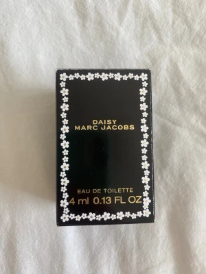 Marc Jacobs Daisy Eau de Toilette 4ml - Söt miniflaska av Daisy från Marc Jacobs, Eau de Toilette 4 ml. Flaskan är genomskinlig med vit blomformad kork och gul detalj. Förpackningen är svart med vita blommor och guldfärgad text. Perfekt att ha i väskan.