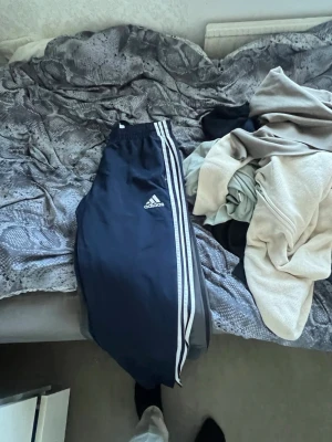 Svarta Adidas mjukisbyxor med vita ränder - Snygga svarta mjukisbyxor från Adidas med klassiska vita ränder längs sidorna och logga på låret. Byxorna har resår i midjan och är perfekta för en avslappnad stil. Passar dig som gillar sportigt mode.