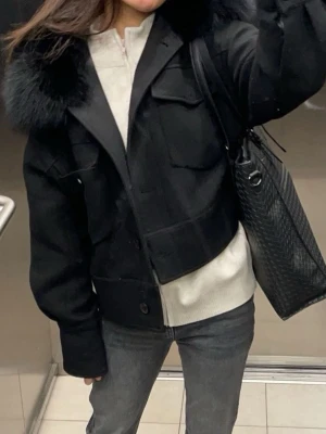 Meotine Charlie jacket - Stilren svart jacka från Meotine med stor krage i fuskpäls. Jackan har två stora bröstfickor med lock och knappar framtill. Kortare modell som ger en trendig siluett. Perfekt för dig som vill ha en snygg och varm jacka med attityd.