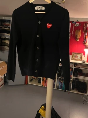 Svart cardigan från Comme des Garçons - Svart cardigan med knappar från Comme des Garçons PLAY. Klassisk design med röd hjärtapplikation med ögon på bröstet. Långärmad och v-ringad, tillverkad i mjukt material som ger en skön känsla. Perfekt för att lyfta din outfit med en ikonisk detalj. Inga deflekter. Tveka inte på att ställa frågor. Priset är diskuterbart vid snabbt köp