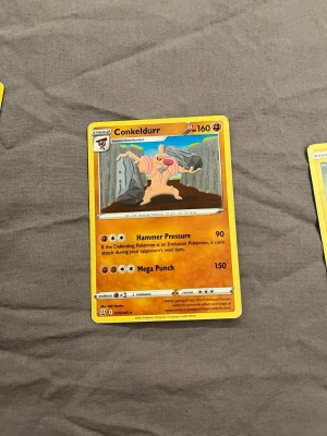 Pokémonkort Conkeldurr - Säljer ett Pokémonkort med Conkeldurr, en fighting-typ med 160 HP. Kortet har en gul ram och motivet visar Conkeldurr med två betongpelare. Text och detaljer i svart, orange och grått. Perfekt för samlare eller Pokémon-fans.