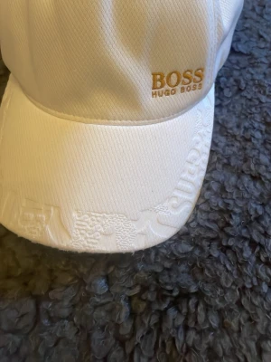 Vit keps från Hugo Boss - Snygg vit keps från Hugo Boss med broderad logga i guld framtill och diskret mönster på skärmen. Klassisk form med böjd skärm och justerbar passform. Tillverkad i ett slitstarkt tyg som känns både stilrent och modernt.