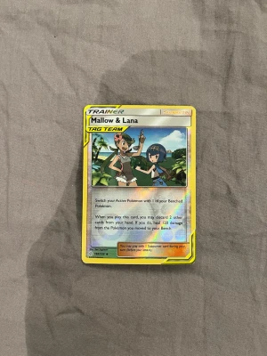 Pokémonkort Mallow & Lana TAG TEAM - Säljer ett Pokémonkort med Mallow & Lana TAG TEAM. Kortet har gul ram och färgglad illustration med två karaktärer i förgrunden. Texten är på engelska och kortet är av typen Trainer Supporter. Perfekt för samlare eller Pokémonfans.