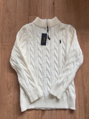 Vit kabelstickad tröja Polo Ralph Lauren - Kabelstickad vit tröja från Polo Ralph Lauren med halv dragkedja och ribbad krage. Tröjan har det klassiska broderade logotypen på bröstet och ribbade muddar vid ärmslut och nederkant. Perfekt för en stilren och avslappnad look.