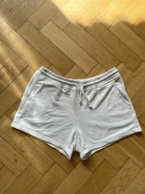 Vita mjuka shorts med snörning - Säljer ett par vita shorts med elastisk midja och snörning framtill. De har två fickor på sidorna och är gjorda i ett mjukt bomullsmaterial. Perfekta för sommardagar eller chill hemma.