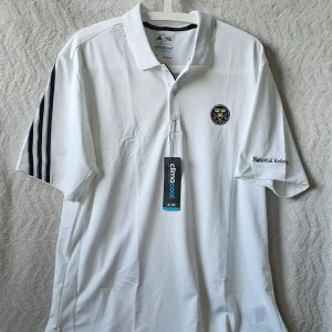 Vit Adidas Climalite pikétröja - Vit pikétröja från Adidas med Climalite-teknologi, svart broderad logga på bröstet och tre svarta ränder på ärmarna. Tröjan har krage, korta ärmar och tryckt text på ena ärmen. Perfekt för sport eller träning.