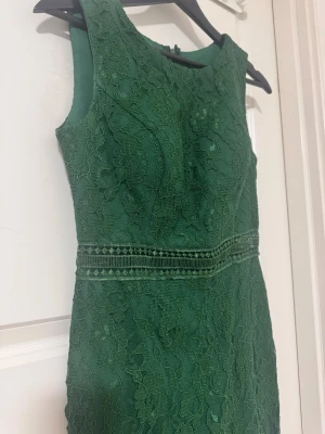 Grön spetsklänning med markerad midja - Snygg grön klänning i spets med ärmlös design och markerad midja. Klänningen har vackra spetsdetaljer och en liten slits baktill. Perfekt för dig som vill sticka ut med en elegant look.