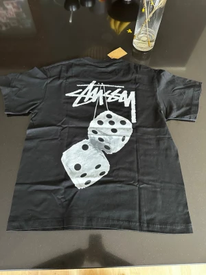 Svart Stüssy t-shirt med tärningsmotiv - Svart t-shirt från Stüssy med stort vitt logotryck och tärningsmotiv på ryggen samt mindre logga på bröstet. Klassisk passform och rund halsringning. Perfekt för dig som gillar streetwear och vill sticka ut med en ikonisk design.