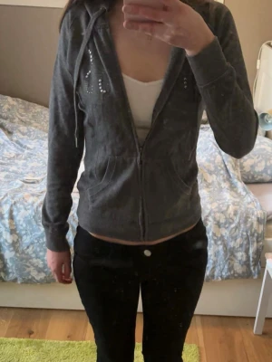 Grå zip-hoodie med strassdetaljer - Mysig grå hoodie från guess med dragkedja, huva och snörning. Framsidan har coola strassdetaljer som bildar text. Perfekt för en chill och avslappnad stil. Två fickor framtill. Materialet känns mjukt och skönt.