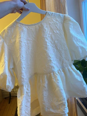 Vit voluminös blus med puffärm - Trendig vit blus med voluminösa puffärmar och rund halsringning. Blusen har en skrynklig textur med blommor och är tillverkad i ett lätt tyg som ger en luftig känsla. Perfekt för att skapa en cool och avslappnad look.