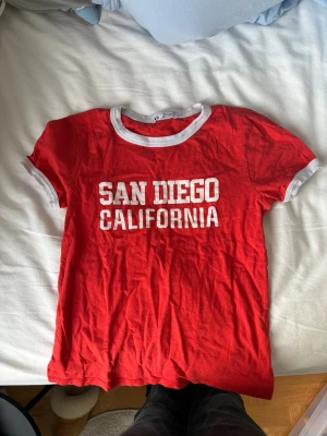 Röd t-shirt San Diego från Zara - Röd t-shirt från Zara med vit text 'San Diego California' på bröstet. T-shirten har vita detaljer vid halsringning och ärmslut. Klassisk passform och korta ärmar, perfekt för en avslappnad stil.