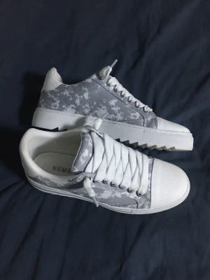 Gråvita sneakers från Numeris - Snygga sneakers i grått och vitt med digitalt camomönster på sidorna. Skorna har vita snören, vit tå och häl samt en robust vit sula med räfflad undersida. Perfekta för dig som gillar en modern och stilren look.