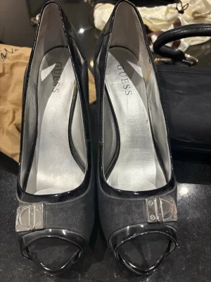 Svarta peep toe pumps från Guess - Snygga svarta pumps från Guess med peep toe och hög klack. Skorna har blanka svarta detaljer och en metallplatta framtill med Guess-logga. Perfekta för dig som vill ha en stilren och trendig look.
