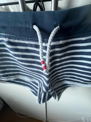 Randiga blåvita shorts med snörning - Säljer ett par blåvita shorts med horisontella ränder och bred midja. Shortsen har vit snörning med röda detaljer och är perfekta för sommaren. De är tillverkade i mjuk bomull och har en sportig, loose passform.