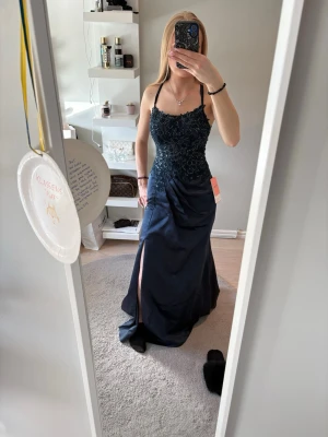 Mörkblå långklänning med spetsdetaljer - Elegant mörkblå långklänning med smala axelband som korsas i ryggen. Klänningen har vacker spets och pärldekor upptill, markerad midja och en slits framtill. Perfekt för bal eller festliga tillfällen.