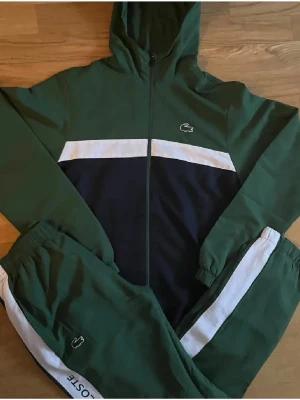 Grön och mörkblå tracksuit från Lacoste - Snygg tracksuit från Lacoste i grönt och mörkblå med vita detaljer. Setet består av en zip-vindjacka med huva och matchande byxor, båda med Lacoste-logga. Materialet är troligtvis polyester, perfekt för sport eller chill. Klassisk streetwear-look med elastiska muddar och dragkedja.