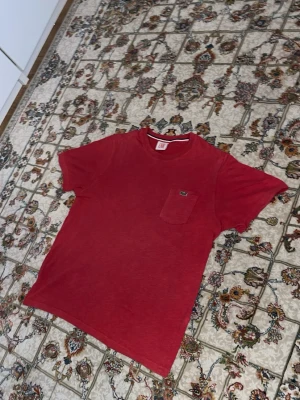 Röd Lacoste LIVE t-shirt med ficka - Snygg röd t-shirt från Lacoste LIVE med klassisk rund hals och en liten bröstficka med den ikoniska krokodilloggan. T-shirten har korta ärmar och är gjord i mjuk bomull, perfekt för en avslappnad och stilren look.