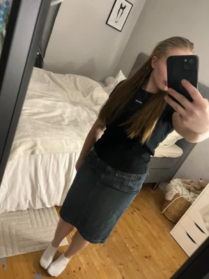 Mörk jeanskjol i klassisk modell - Säljer en mörkblå jeanskjol i knälång modell med klassiska fickor och bälteshällor. Kjolen har en rak passform och är perfekt för dig som gillar en enkel och tidlös stil. Passar till både t-shirt och stickad tröja.