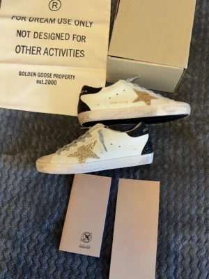 Golden Goose sneakers med glitterstjärna - Snygga vita Golden Goose sneakers med en glittrig guldstjärna på sidan och svart glitter på hälen. Skorna har grå skosnören och klassisk låg modell. Tillverkade i läder med detaljer i glitter för en unik look.