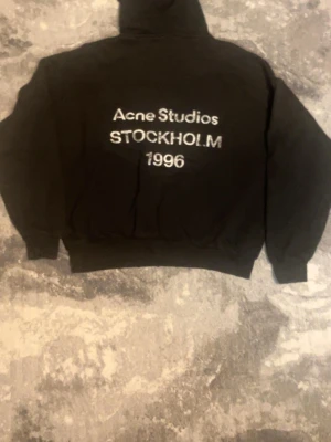 Svart hoodie Acne Studios Stockholm - Svart hoodie från Acne Studios med vit text på ryggen där det står 'Acne Studios STOCKHOLM 1996'. Klassisk passform med huva och ribbade muddar. Perfekt för en stilren och trendig look.