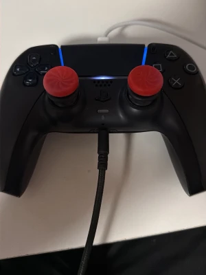 Vit  PS5 DualSense handkontroll - Säljer en svart PlayStation 5 DualSense handkontroll med röda joystick-skydd. Kontrollen har touchpad, LED-ljus och klassiska PlayStation-knappar. Ergonomisk design i plast och ansluts via USB-C. Perfekt för gaming på PS5. Har stick drift 