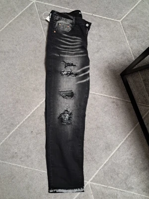 Svarta ripped jeans från Purple Brand - Säljer ett par svarta jeans från Purple Brand med snygga slitningar och trasiga detaljer på framsidan. Jeansen har klassisk femficksdesign och smal passform. Perfekta för dig som gillar en edgy och trendig stil.