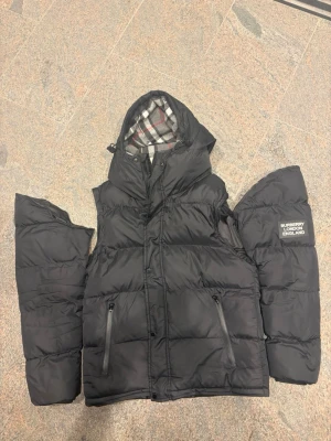 Svart pufferjacka från Burberry - Snygg svart pufferjacka från Burberry med avtagbara ärmar och huva. Jackan har två dragkedjefickor framtill och klassiskt rutigt foder i huvan. Märkeslogga på vänster ärm. Perfekt för kalla vinterdagar.