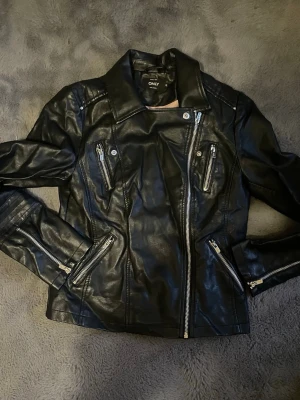 Svart skinnjacka från ONLY - Snygg svart skinnjacka från ONLY med coola detaljer som snedställd dragkedja, fyra dragkedjeförsedda fickor och markerade axlar. Jackan har en klassisk bikerstil och är perfekt för dig som vill ha en edgy look. Materialet är skinn och jackan har en figurnära passform. Obs går att upphämtas i södra Stockholm !!!