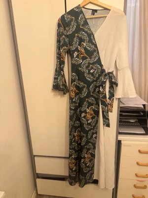 Omlottklänning med blommigt mönster - Unik lång omlottklänning med ena sidan i beige och andra sidan i grönt med blommigt mönster. Klänningen har långa trumpetärmar och knyt i midjan för en snygg siluett. Perfekt för dig som vill sticka ut med en cool och trendig look.