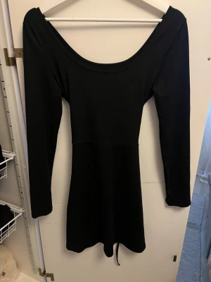 Svart långärmad kortklänning - En svart, långärmad kortklänning med urringning och korsettliknande snörning i ryggen. Klänningen har en enkel, stilren design och är tillverkad i ett mjukt, stretchigt material som ger en bekväm passform.
