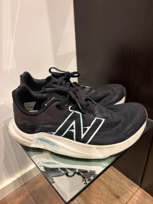 Svarta New Balance sneakers - Snygga svarta sneakers från New Balance med vit sula och stor vit N-logga på sidan. Ovandelen är i meshmaterial för bra andningsförmåga och skorna har klassisk snörning. Perfekta för dig som gillar sportig och stilren look.