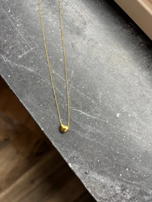 Edblad halsband - Ett elegant halsband i guldton med en liten hjärtformad berlock. Kedjan är tunn och stilren, perfekt för att ge en subtil touch till din look. Materialet ser ut att vara metall med guldfinish och berlocken har en mjuk, rundad form.